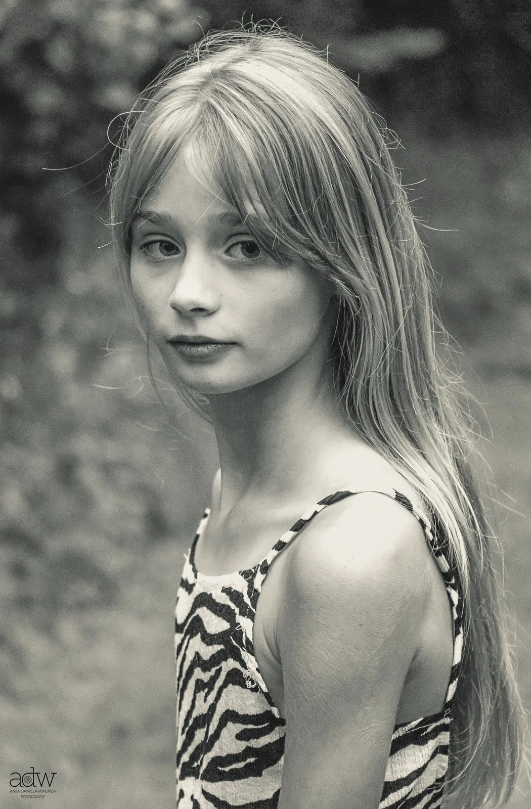Black and white portrait of a young woman with long blonde hair, wearing a sleeveless zebra-print top, looking over her shoulder outdoors.