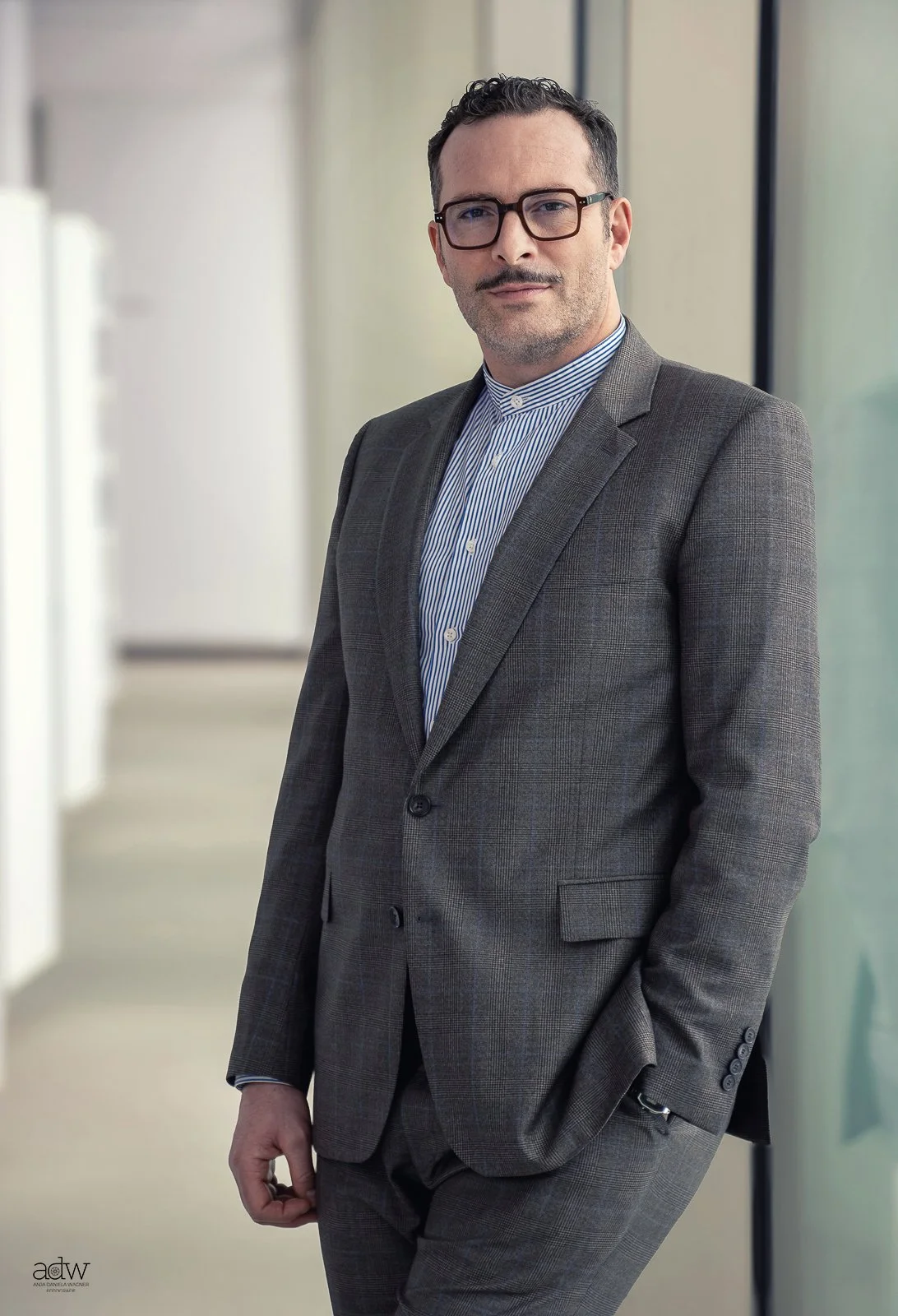 A man in a gray plaid suit with a striped shirt, wearing glasses, standing in a modern indoor setting with large windows.