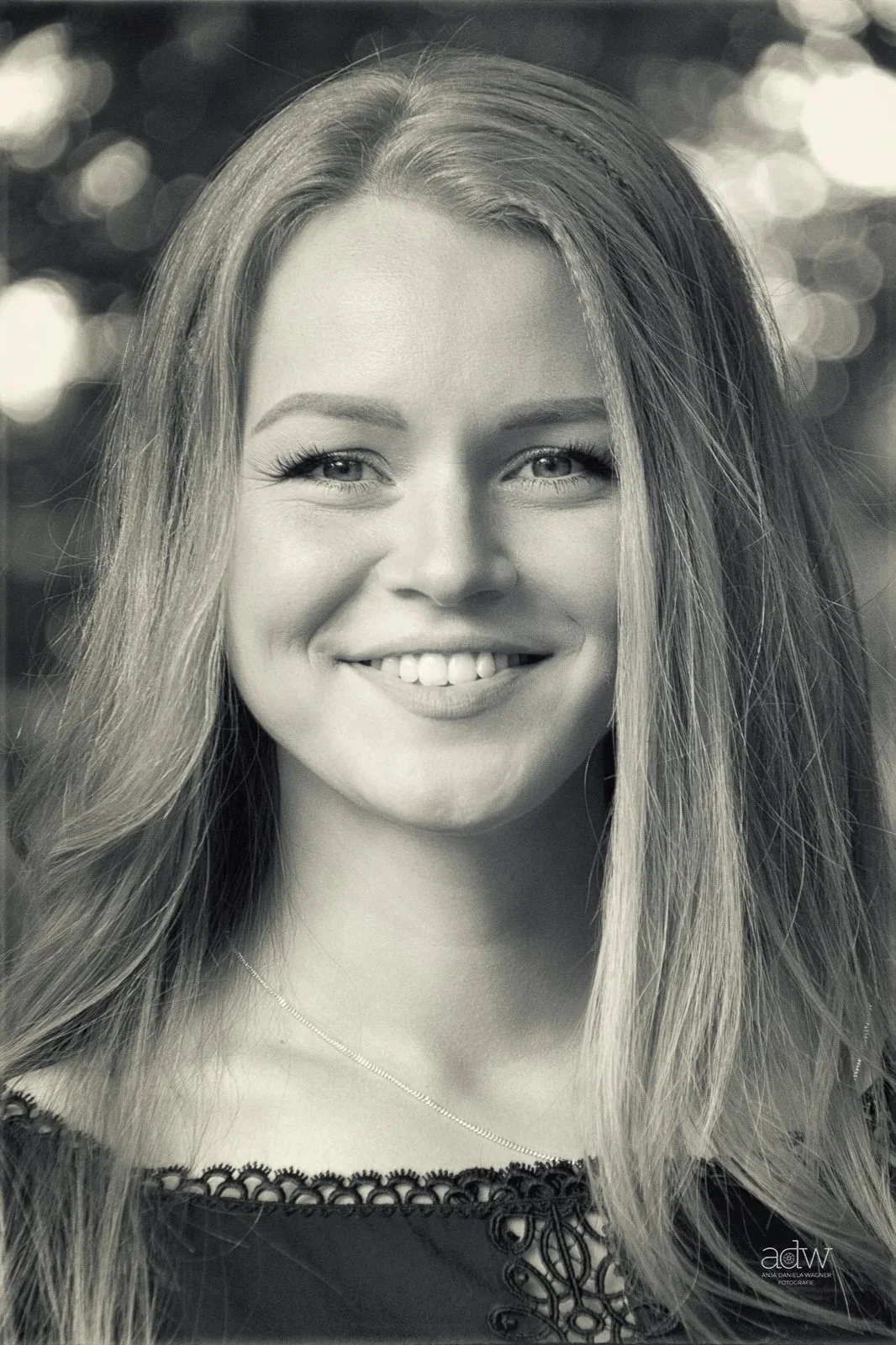 Black and white portrait of a young woman with long, wavy hair, smiling, wearing a necklace, in front of gossamer lights.