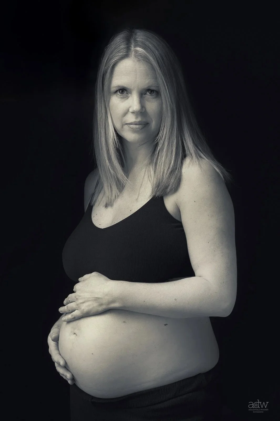 Black and white portrait of a pregnant woman with long hair wearing a black tank top, looking at the camera, holding her belly.