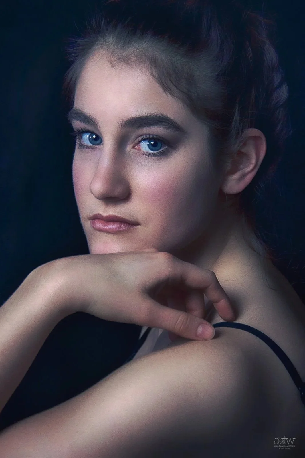 Close-up portrait of a young woman with blue eyes and dark hair, resting her hand on her shoulder, against a dark background.