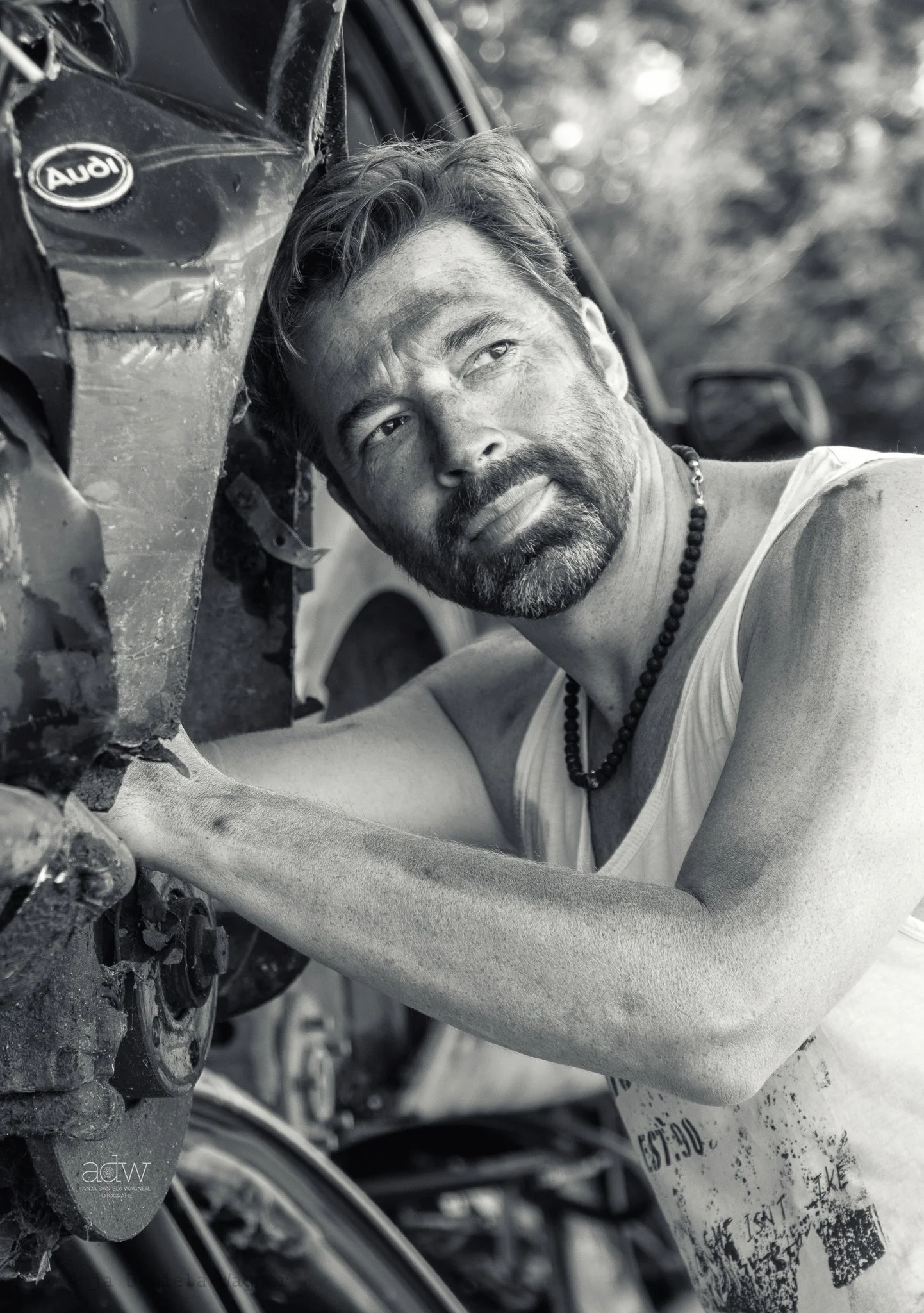 Close-up of a person's face, lying next to a damaged motorcycle with visible dirt, mud, and mud. The person is gazing into the distance with a calm expression and wearing a beaded necklace and a sleeveless shirt.