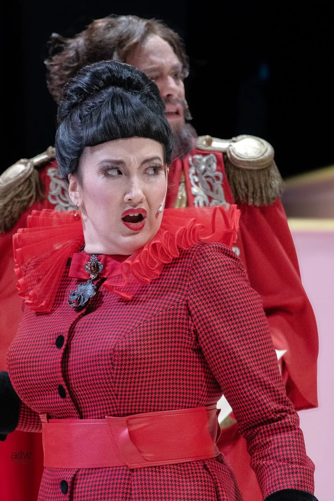 A woman dressed in a red checkered dress with a ruffled collar and shoulder epaulets, making a surprised or shocked facial expression, with a man dressed in a red military uniform with gold epaulets in the background.