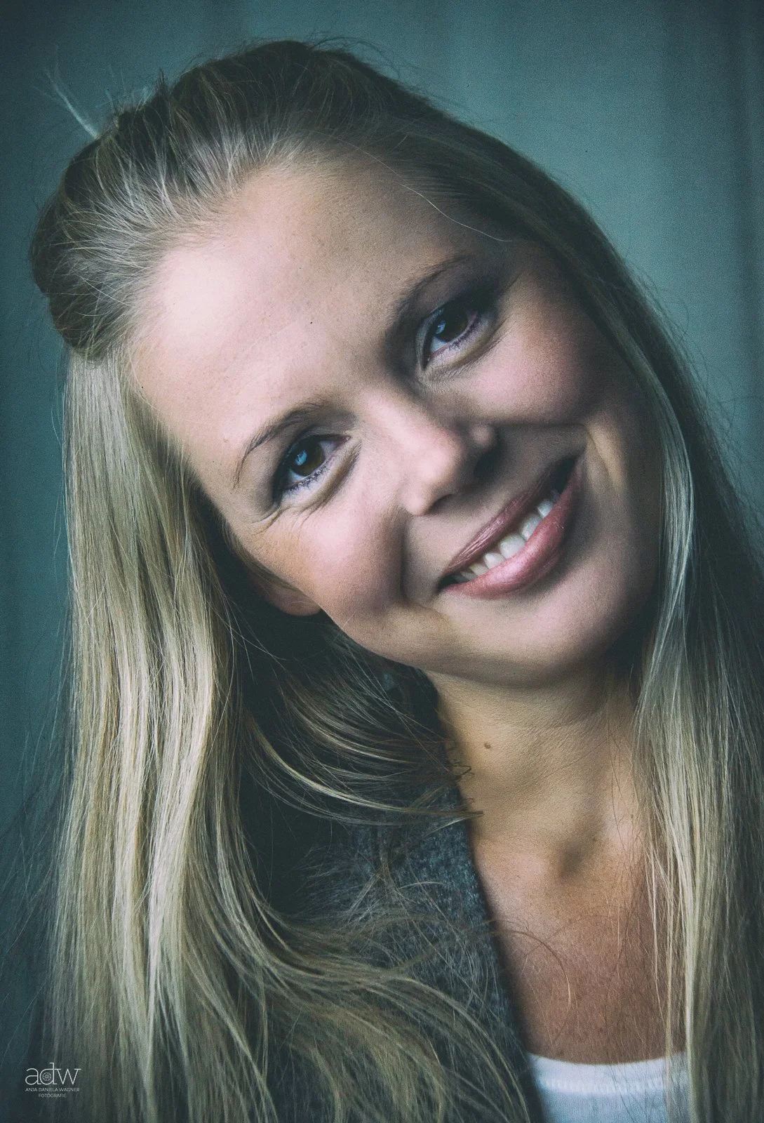 Close-up portrait of a smiling woman with long blond hair, blue eyes, and light makeup, wearing a gray jacket over a white top.