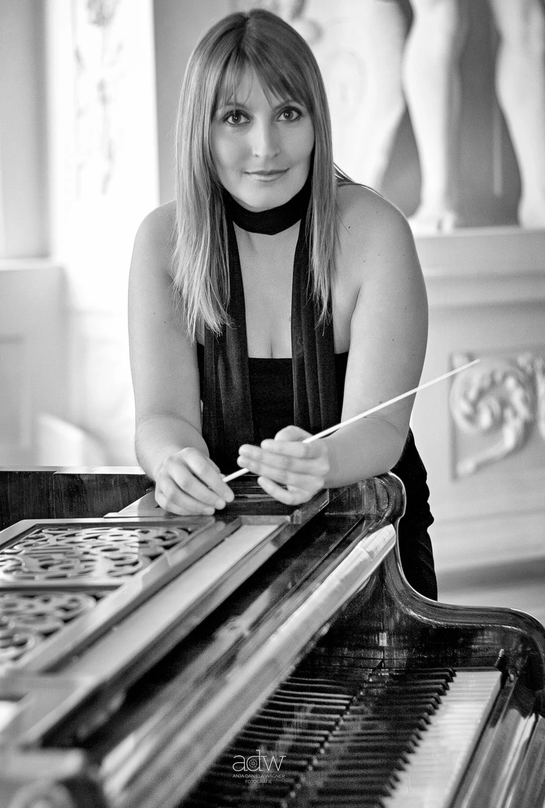 A woman with shoulder-length hair and a black sleeveless top is sitting at a vintage piano, holding a conductor's baton, in a room with classical sculptures.
