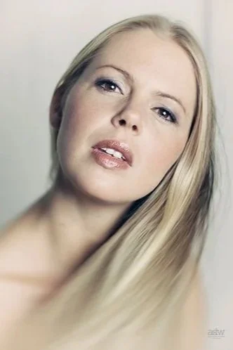 A close-up portrait of a blonde woman with light makeup, slightly parted lips, and blue eyes looking at the camera.