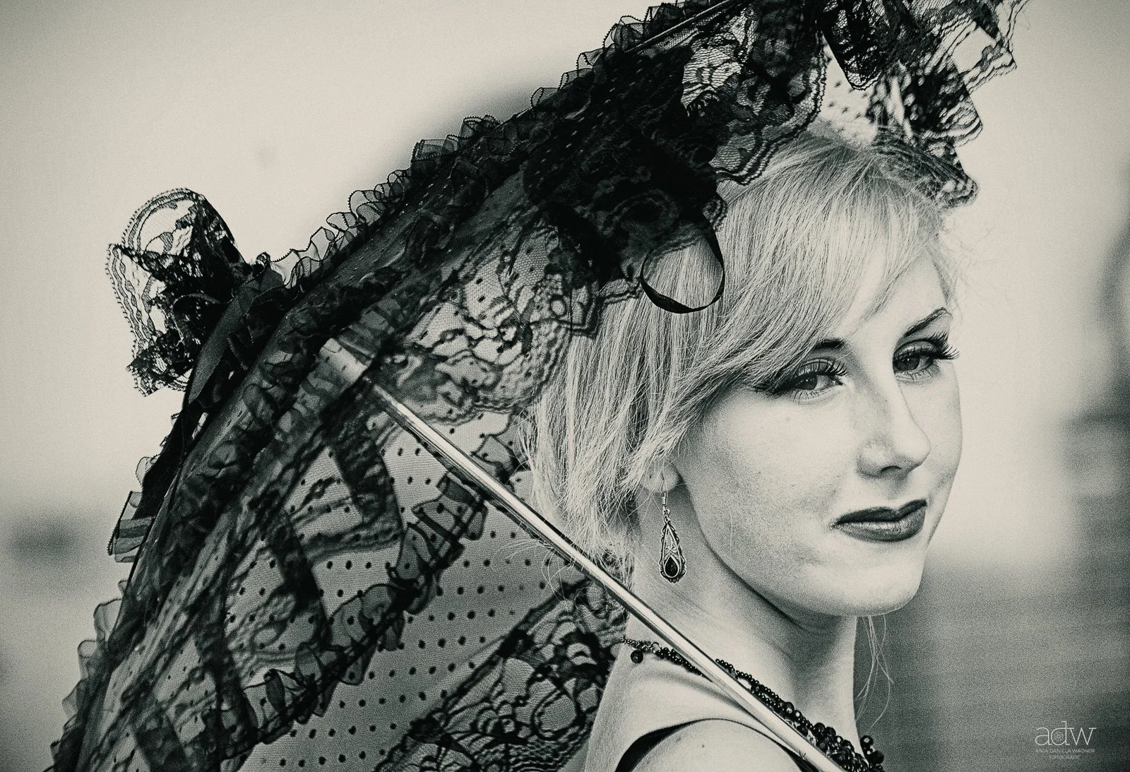 Black and white portrait of a woman with short blonde hair, wearing lace clothing and earrings, holding a lace parasol.