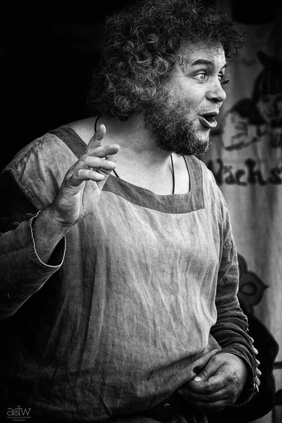 A man with curly hair and a beard, wearing a loose, long-sleeve shirt, is speaking and gesturing with his hand, in black and white.