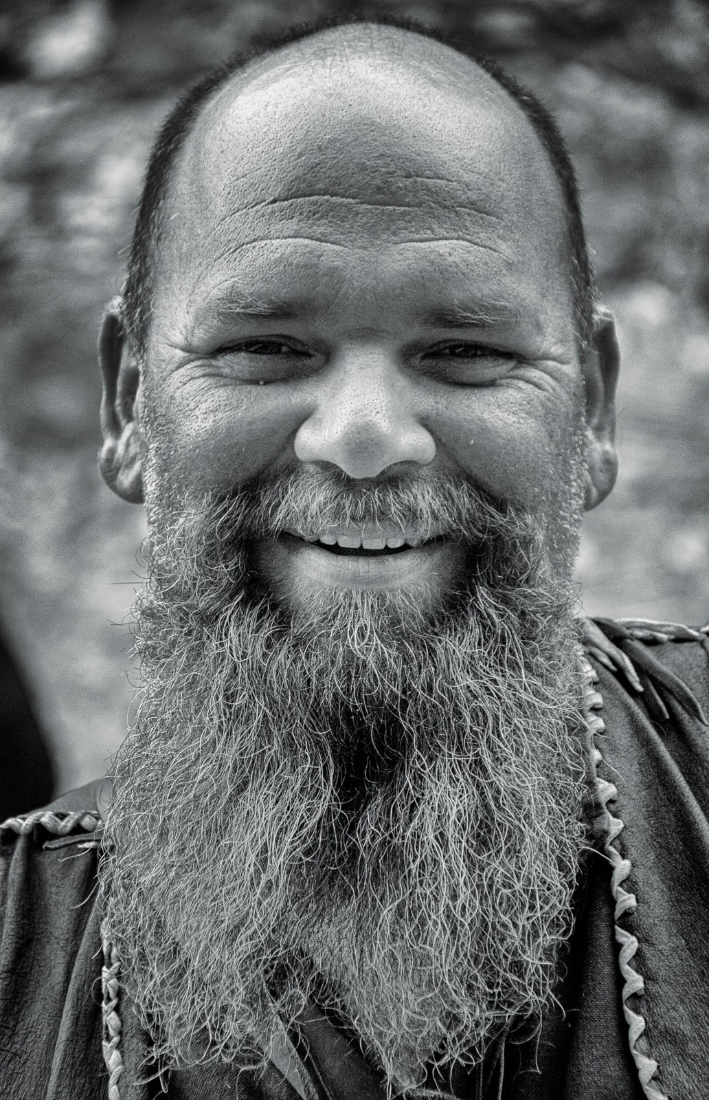 A smiling man with a shaved head and a long, thick beard, wearing a dark shirt with decorative stitching on the sleeves, outdoors.