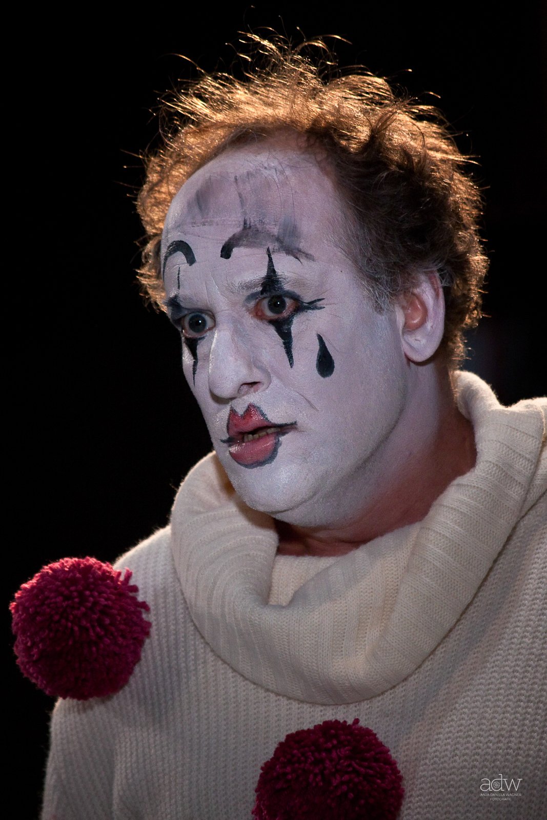 A person dressed as a clown with white face paint, black makeup accents, and a red nose, speaking into two red microphones.