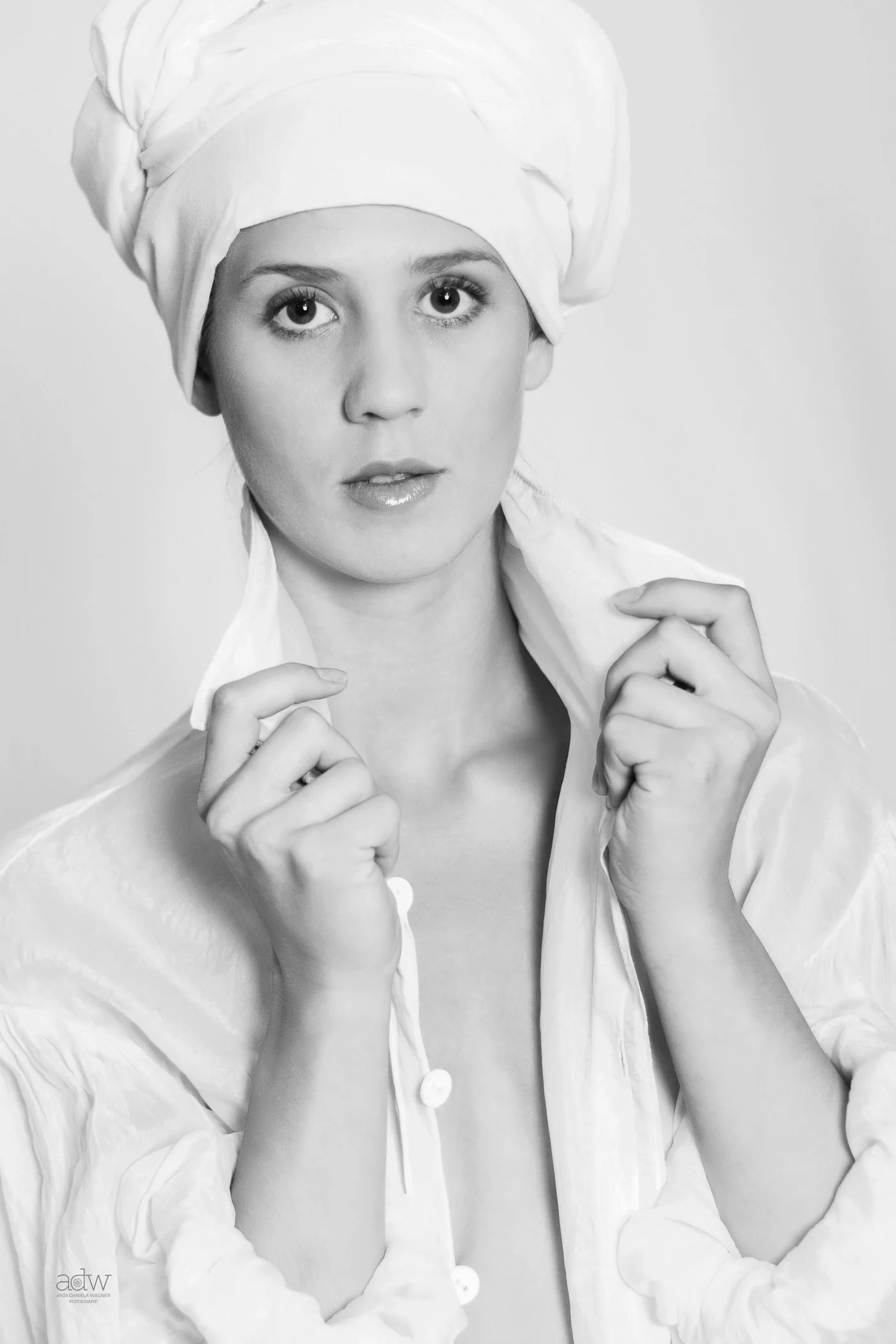 A black and white portrait of a woman with large eyes, wearing a white headwrap and a light-colored silk shirt, holding the shirt's collar with both hands.