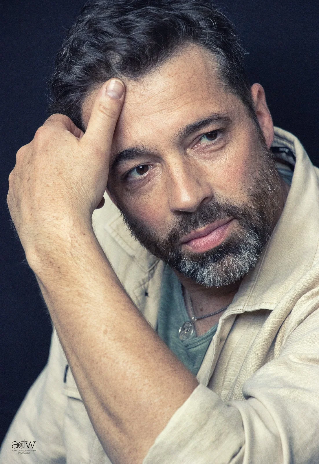 A man with dark curly hair, a beard, and light skin, resting his head on his left hand against a dark background.
