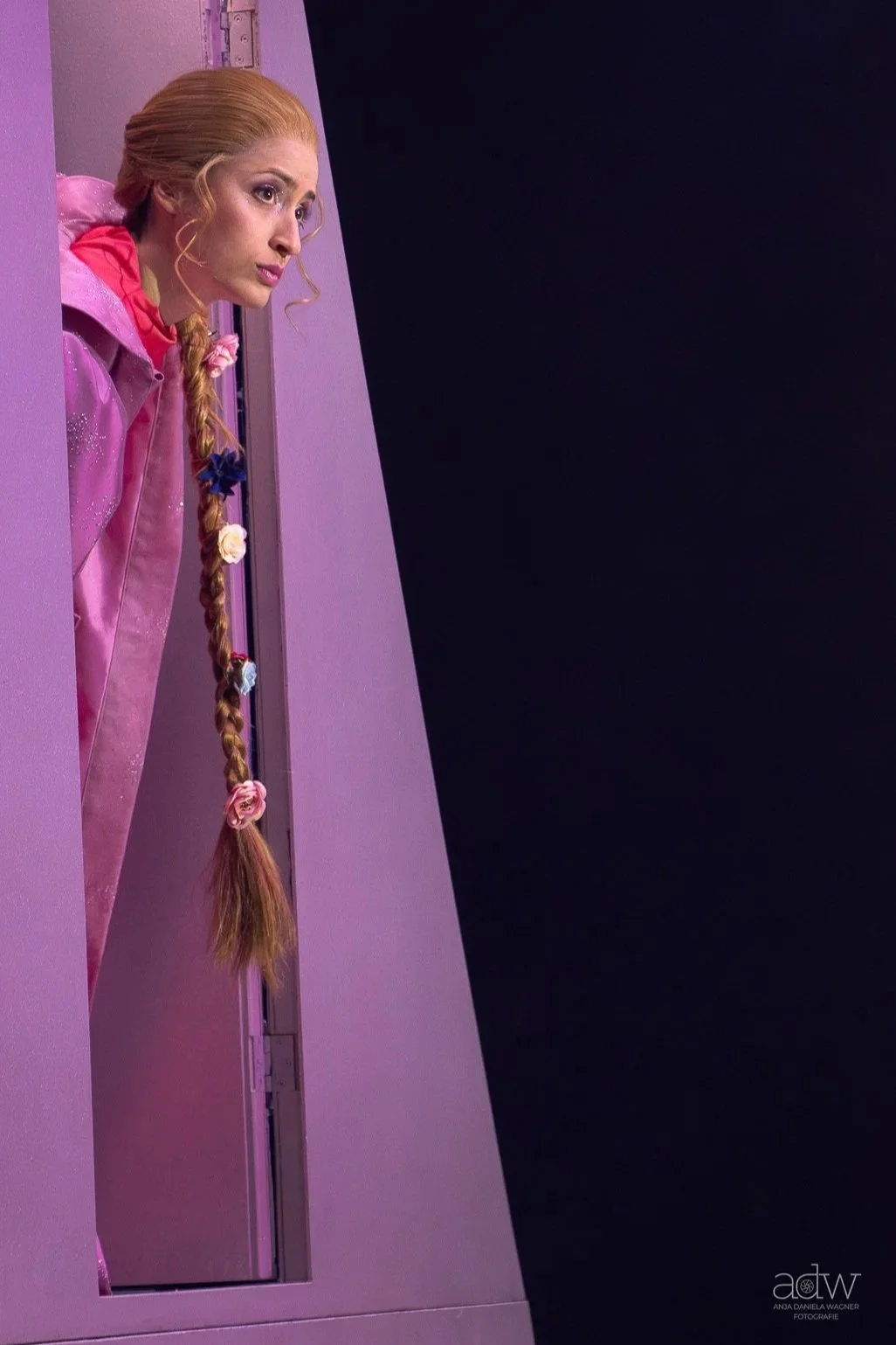 A woman with long, braided hair decorated with colorful flower hair ties, looking out a small window on a pink wall. She is wearing a pink outfit, and the background is dark black.