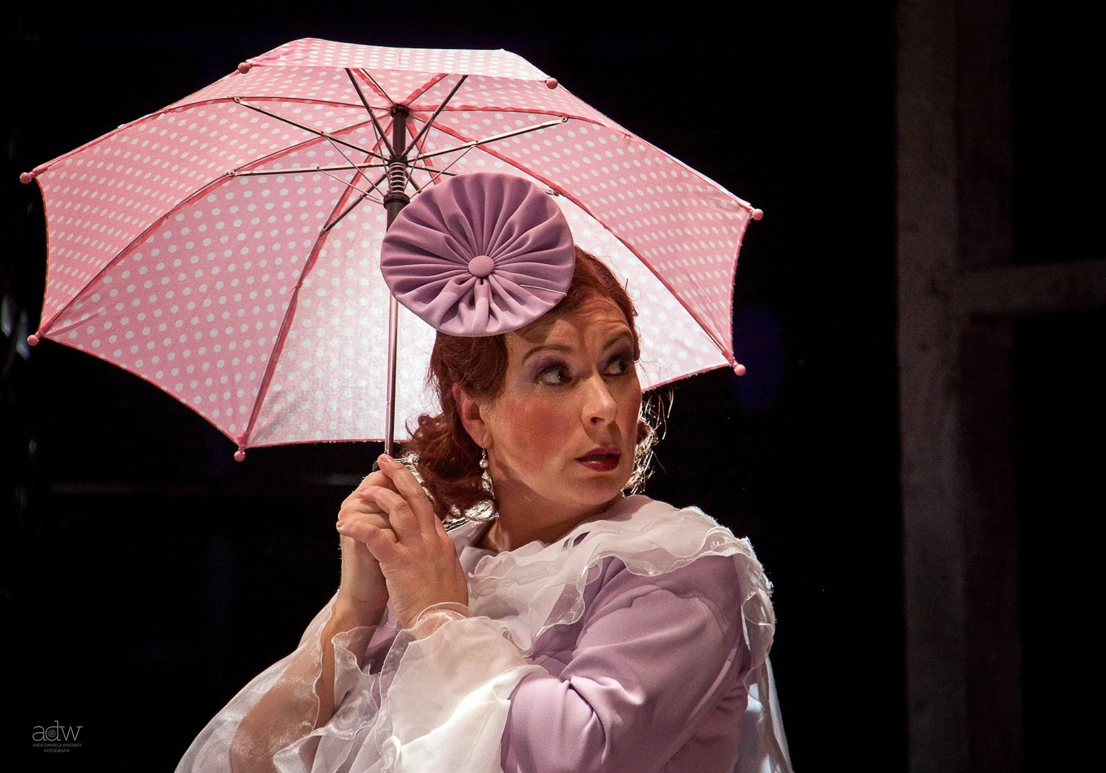 A woman with red hair holding a pink polka dot umbrella. She is looking to her right with a surprised or intrigued expression, wearing a white, ruffled blouse, and a pink headpiece.