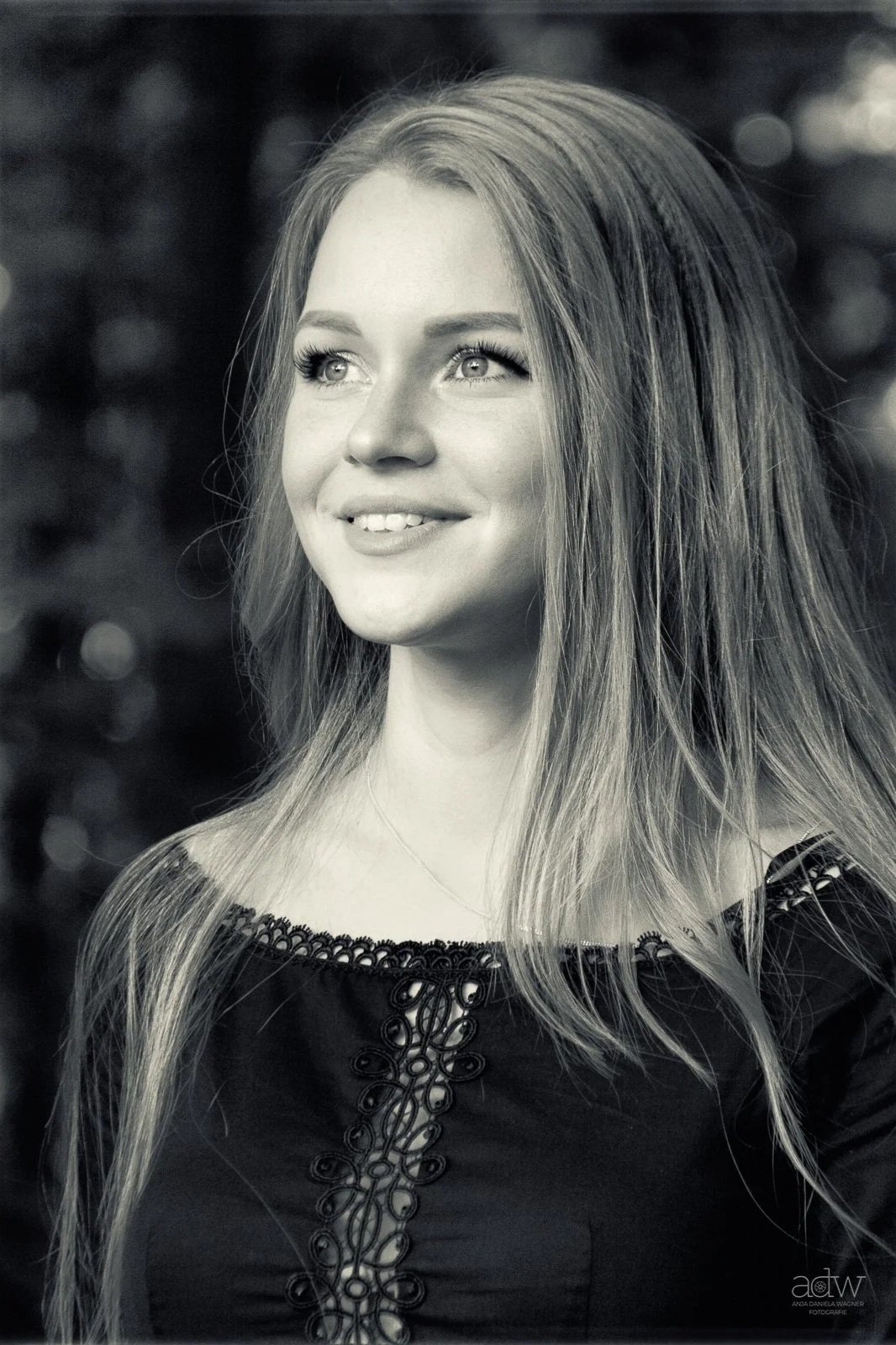A young woman with long, light-colored hair smiling and looking slightly upward. She is wearing a dark top with decorative embroidery at the neckline, and the background is blurred.