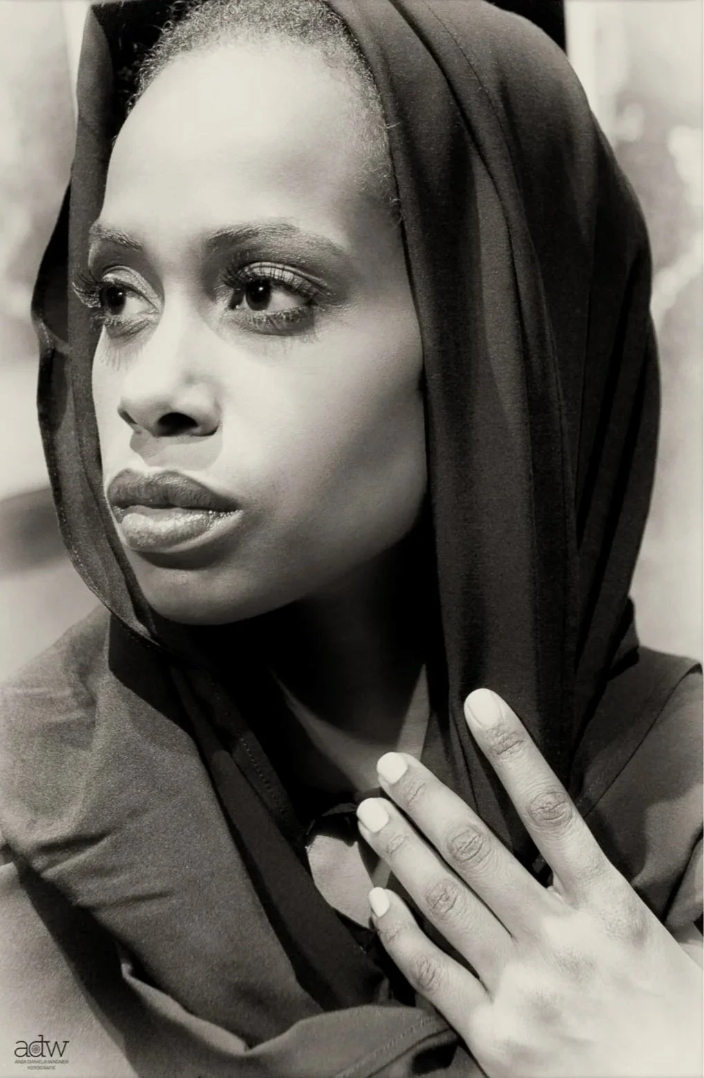 Close-up black-and-white portrait of a woman wearing a headscarf, with her hand gently resting on her chest, showing detailed facial features and expression.