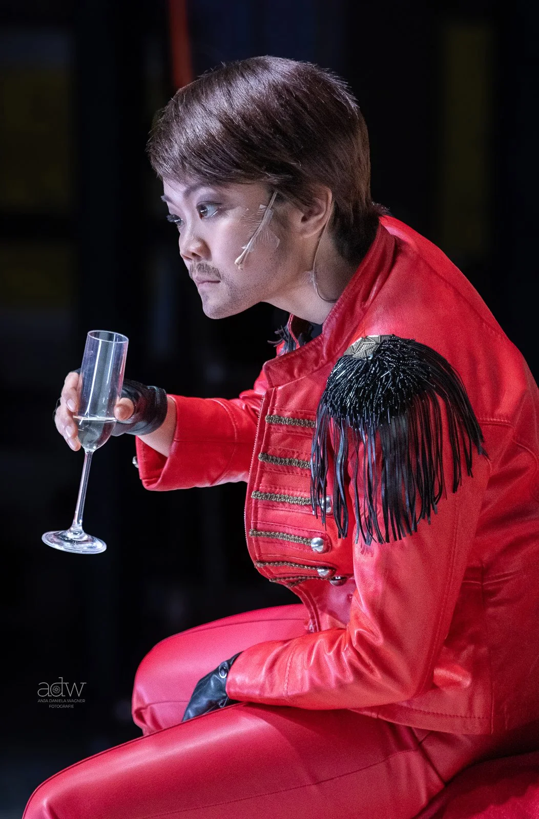 A man dressed in a red military-style jacket with black fringe on the shoulder, holding a glass of champagne, sitting with a contemplative expression in a dimly lit setting.