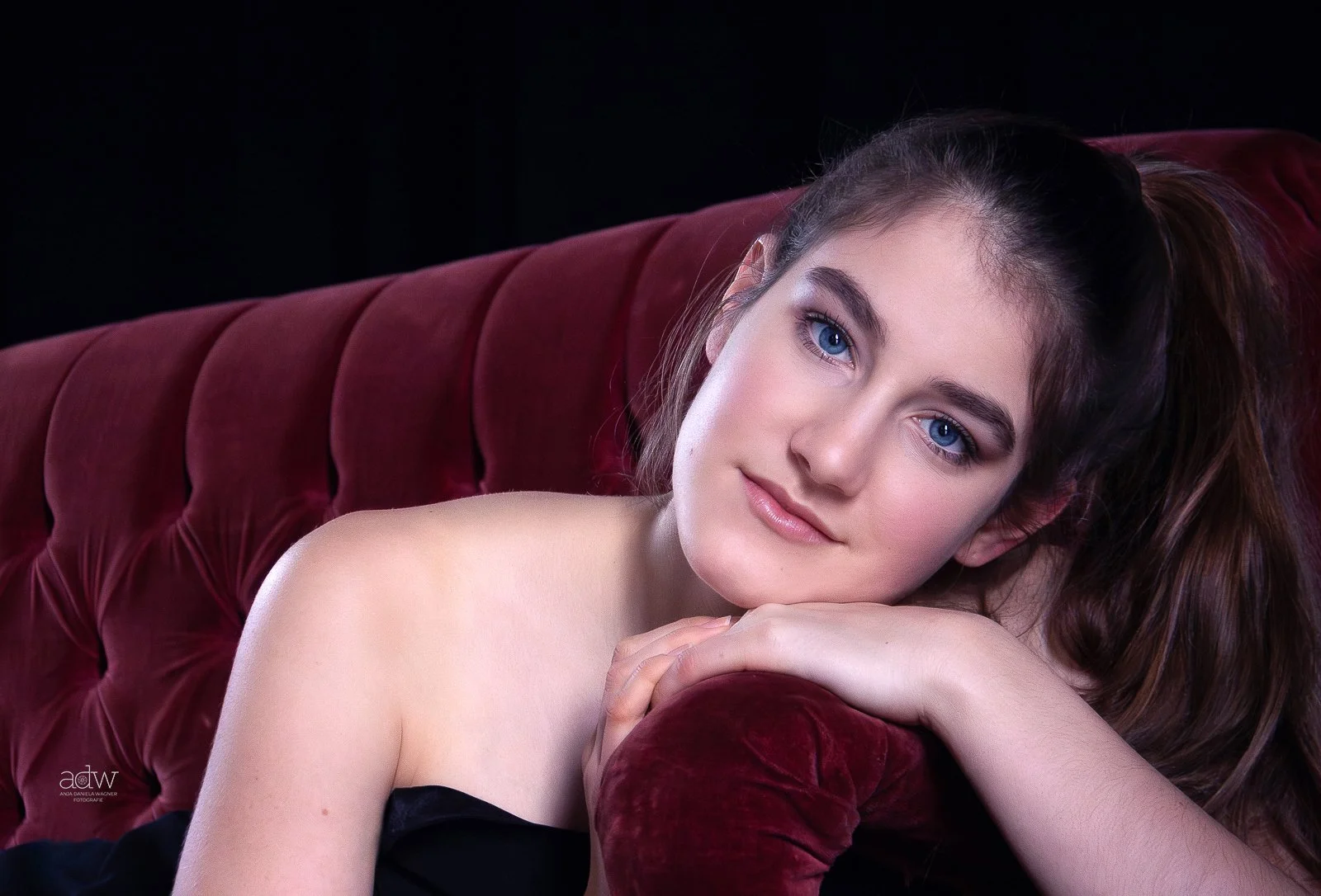 Young woman with blue eyes and brown hair relaxing on a red velvet couch, resting her head on her hands.