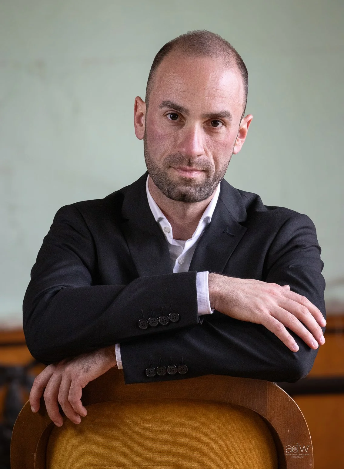 A man with a bald head and beard in a black suit and white shirt, sitting with arms crossed on the back of a wooden chair, in front of a plain green wall.