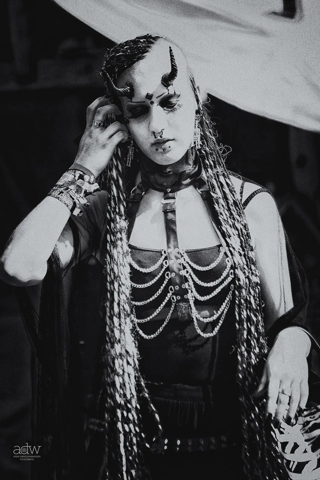 A person with elaborate gothic and punk fashion, including multiple chains, layered jewelry, and detailed makeup, standing with eyes closed and hand touching their face.