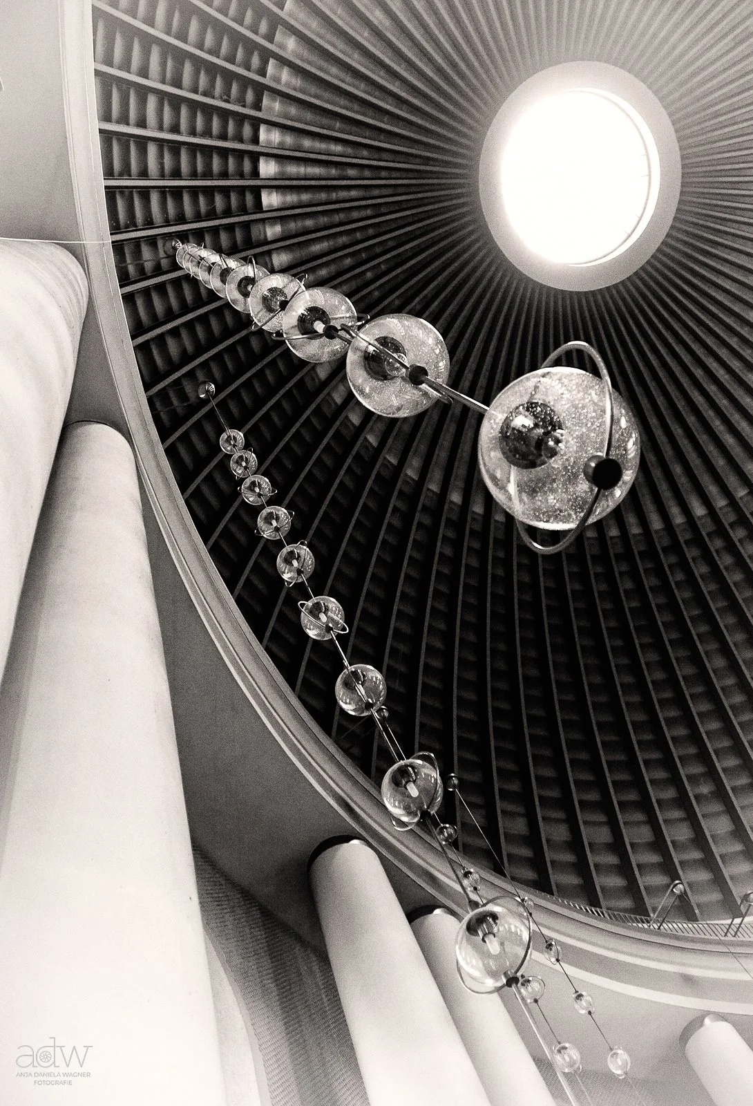 Looking up at a circular skylight in a domed ceiling, with hanging spherical light fixtures and columns supporting the structure.