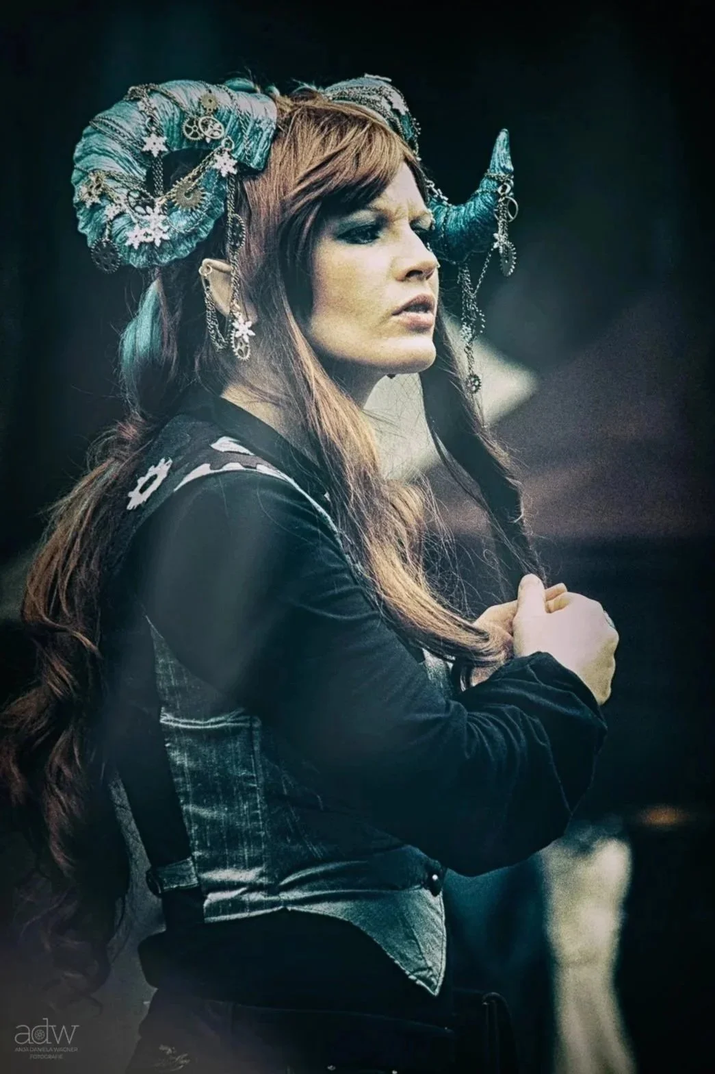 A woman with long brown hair wearing a dark jacket and a headpiece with blue horns decorated with chains and jewelry.