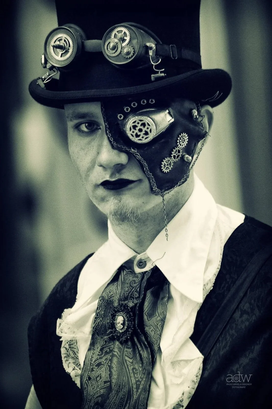 A person wearing a steampunk-inspired costume, including a hat with mechanical gears and goggles, a leather eyepatch with gears, a white shirt, a patterned tie, and dark lipstick, posing in black and white.