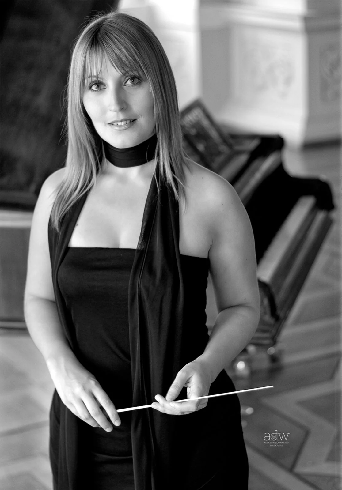 Black and white photo of a woman in formal attire, holding a baton, standing in front of a grand piano in a music room.