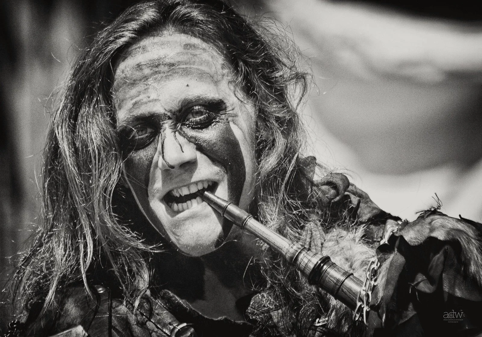 Black and white photo of a person with long hair, face painted in a clown-like manner, playing a flute, wearing a costume with chains and tattered fabric.