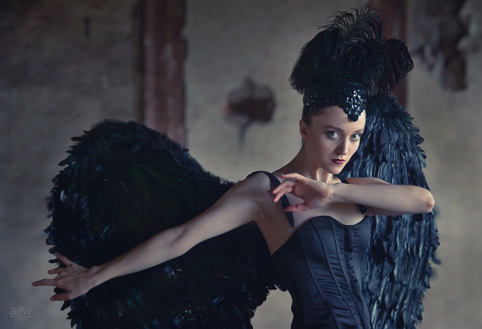A woman in black attire and elaborate feathered headpiece and wings, posing with her arm extended in a dramatic fashion.