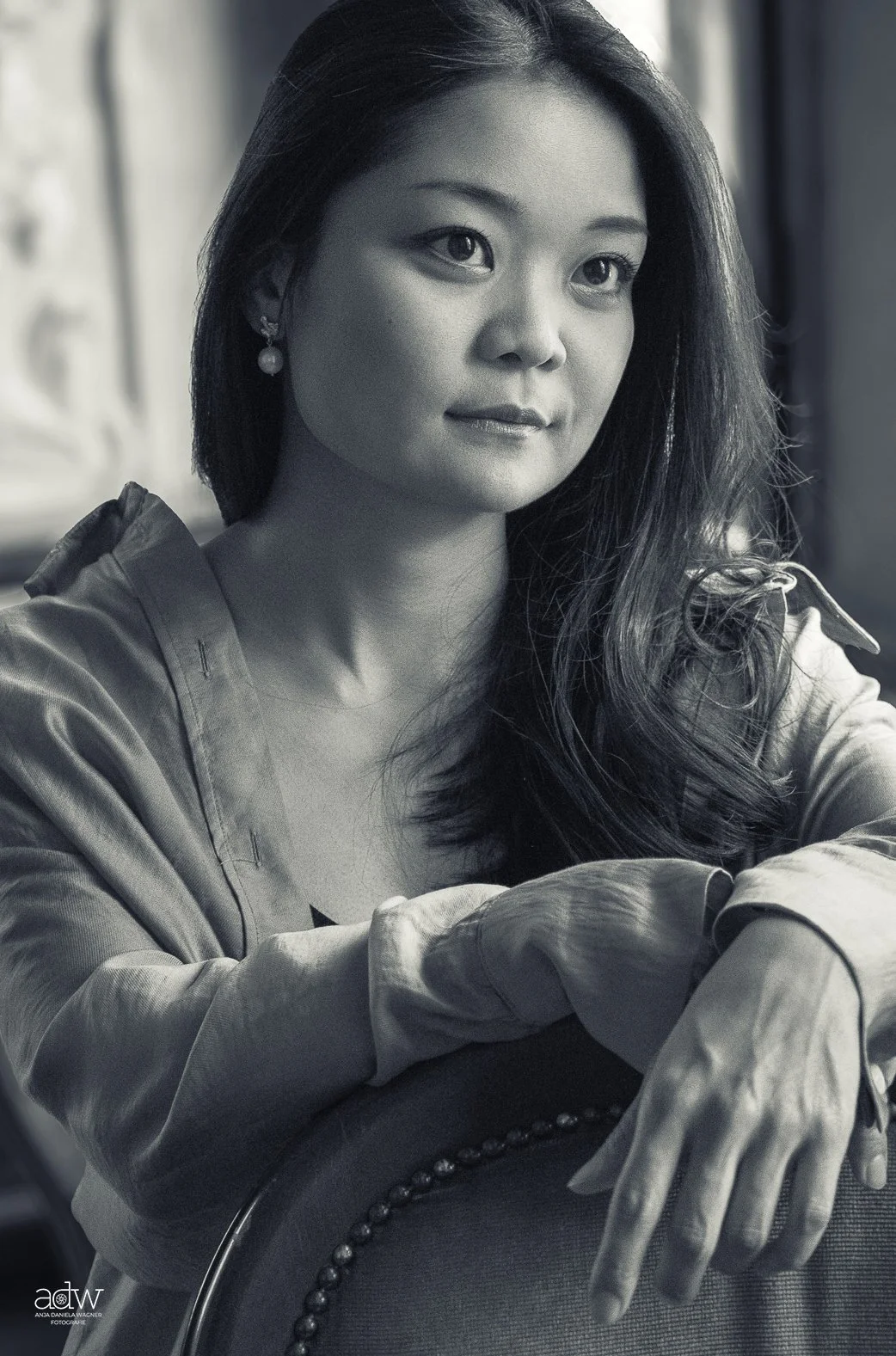 Black and white portrait of a woman with long wavy hair, wearing pearl earrings and a jacket, sitting with arms resting on a chair, looking to the side.