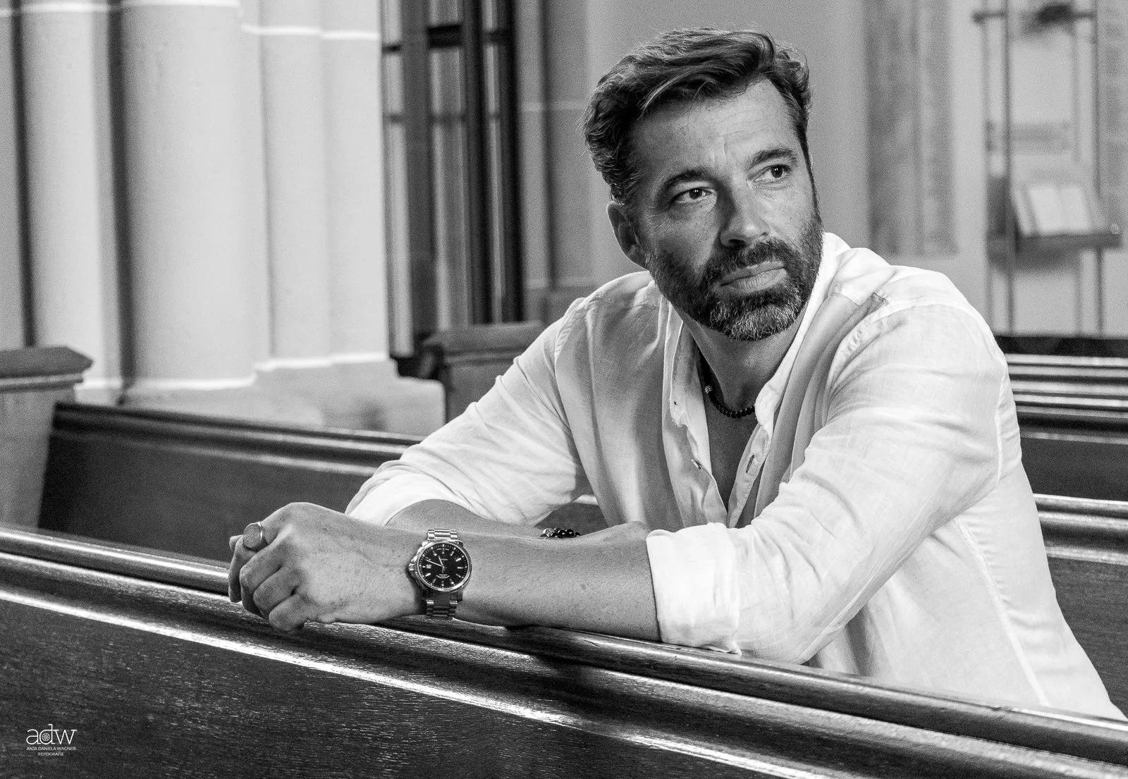 A black-and-white photo of a man with a beard and mustache, sitting at a bench in what appears to be a church or chapel. The man is wearing a white shirt, a watch on his wrist, and a necklace. He is looking to the side with a serious or contemplative expression.