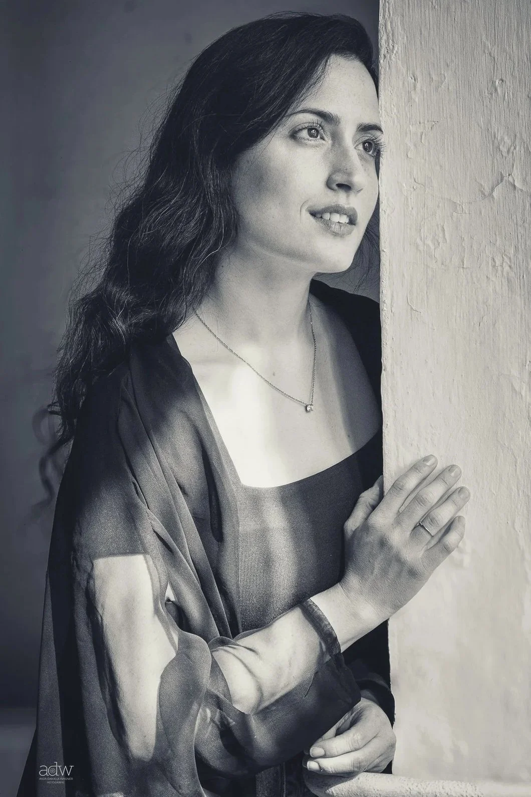 A black and white photo of a woman with dark, wavy hair, looking thoughtfully to the side while holding a textured wall with one hand. She wears a necklace, a dark top, and a sheer, patterned shawl.