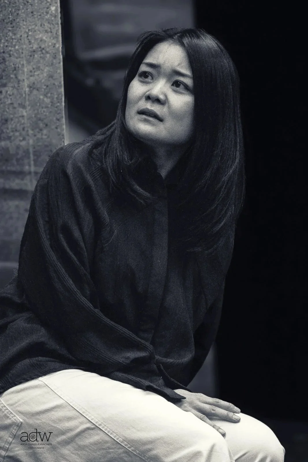 Black and white photo of woman with medium-length dark hair, sitting against a wall, looking upwards with a concerned or thoughtful expression.