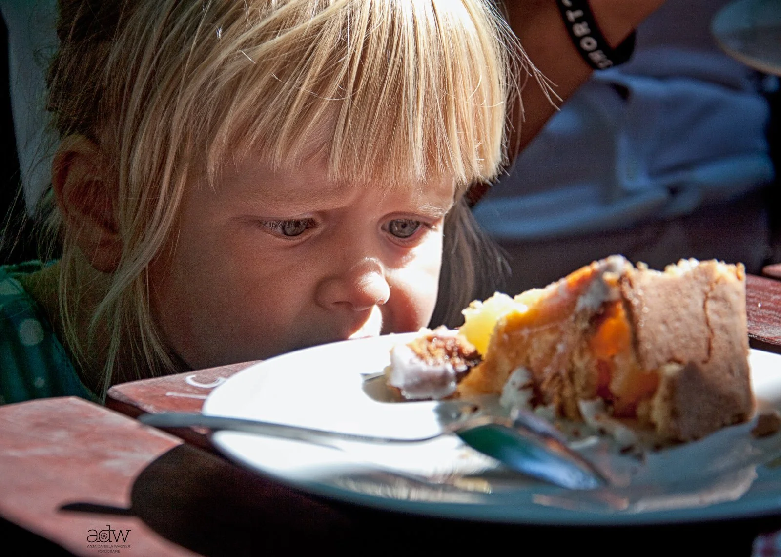 A young girl with blonde hair looking closely at a slice of cake on a plate in front of her.