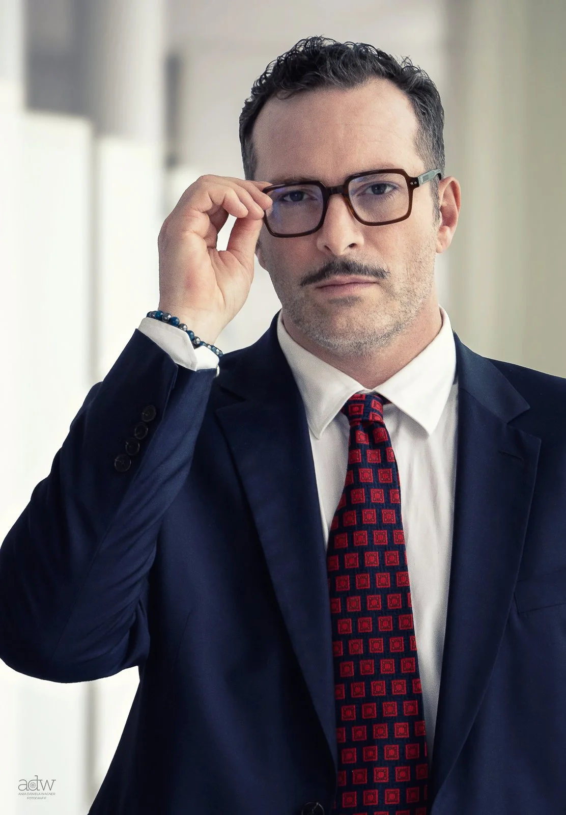 A man in a suit adjusts his glasses in an office setting.