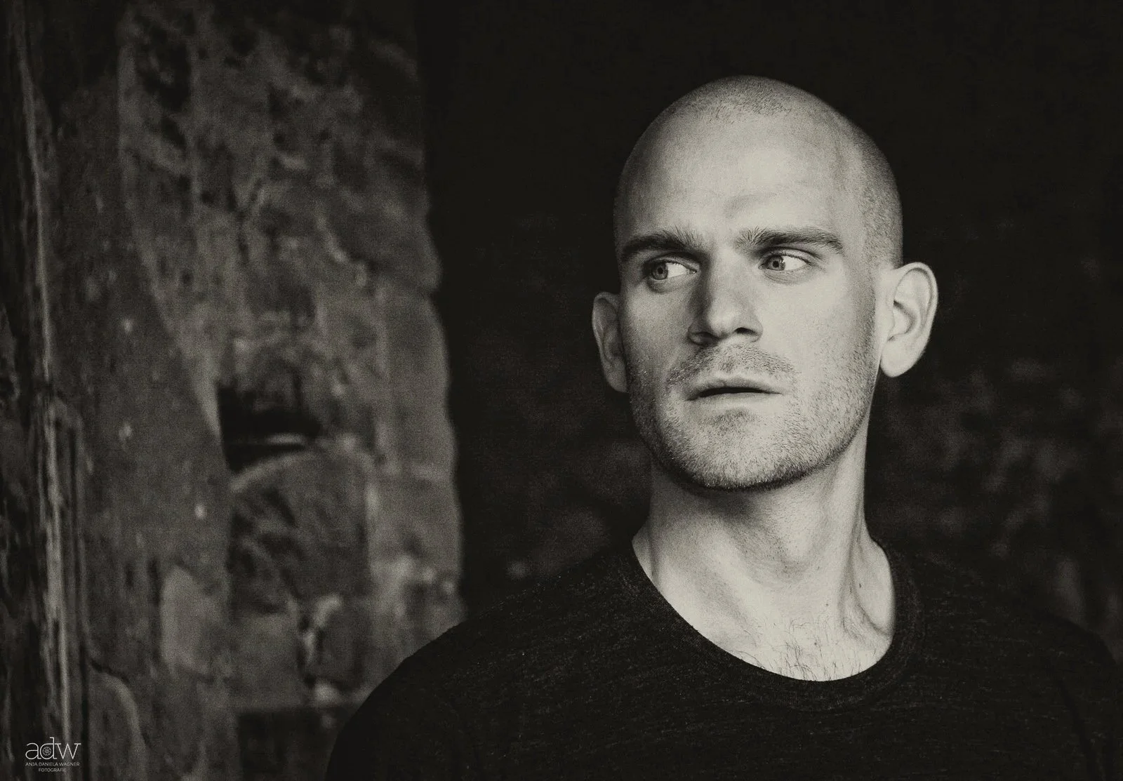 Black and white portrait of a man with a shaved head, looking to the right, next to a brick wall.