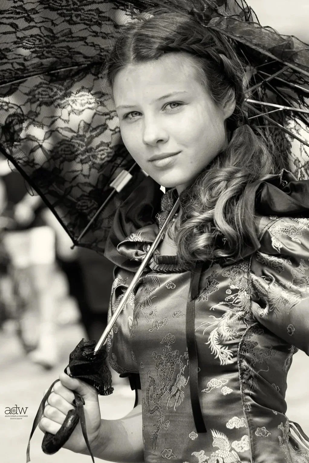 A woman holding a lace umbrella, dressed in a traditional patterned silk garment with Asian dragon designs, smiling softly.