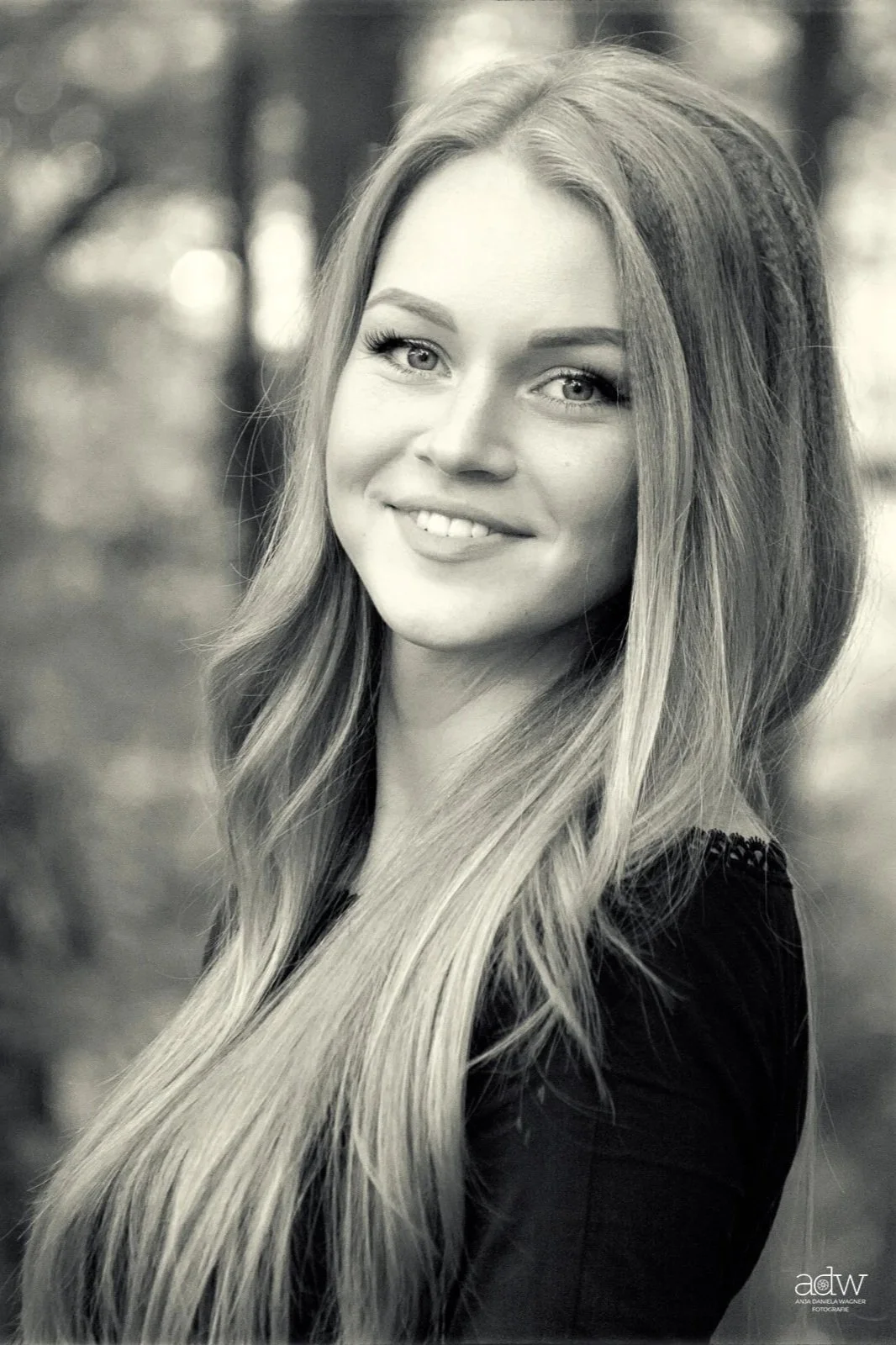 Black and white portrait of a smiling young woman with long, wavy hair, looking at the camera. Blurred outdoor background.