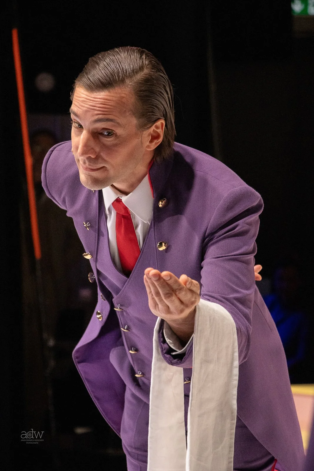 Man in a purple uniform with shiny buttons, white shirt, and red tie, leaning forward and extending one hand as if presenting or offering something.