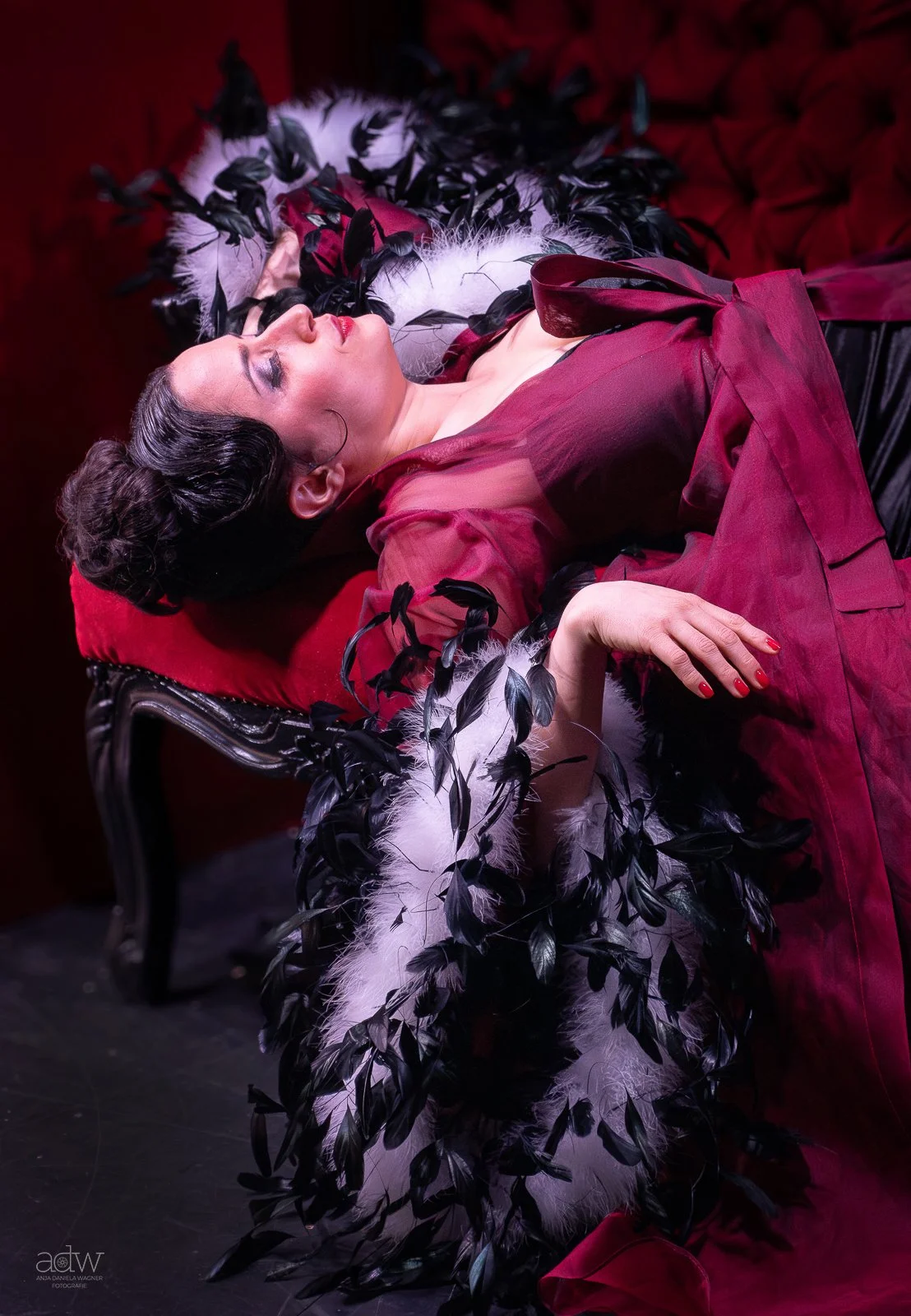 A woman lying on a black ornate chair, dressed in a red dress with sheer sleeves, surrounded by black and white feathered and leaf-like accessories. She has dark hair styled in an elegant updo and is wearing makeup, with her eyes closed and red lipstick.