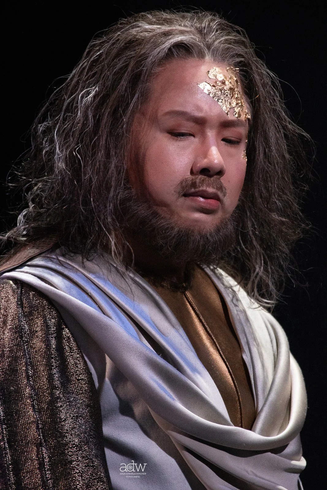 A person dressed as a sci-fi character with long, curly, gray hair, touching gold paint on their forehead, in a costume with metallic and white fabric, standing against a black background.
