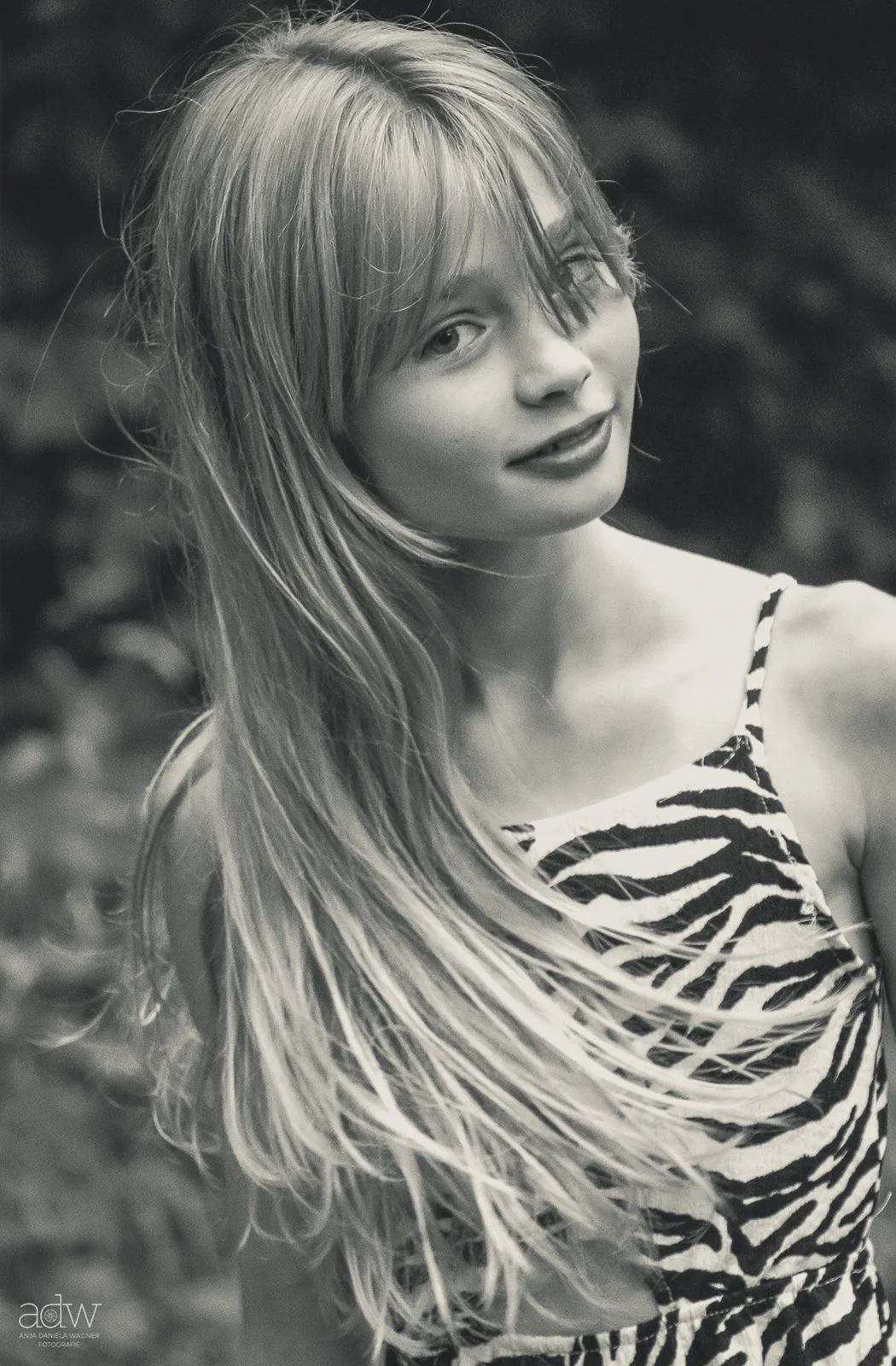Black and white photo of a young woman with long, flowing hair, wearing a zebra print top, standing outdoors with a blurred natural background, smiling slightly.