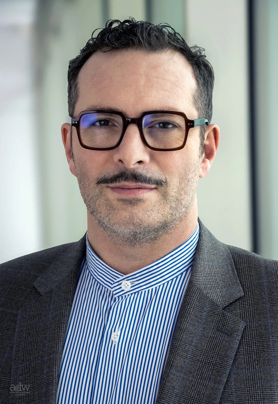 A man with short dark hair, glasses, and a well-groomed beard and mustache, wearing a suit and blue striped dress shirt, standing indoors with a blurred background.