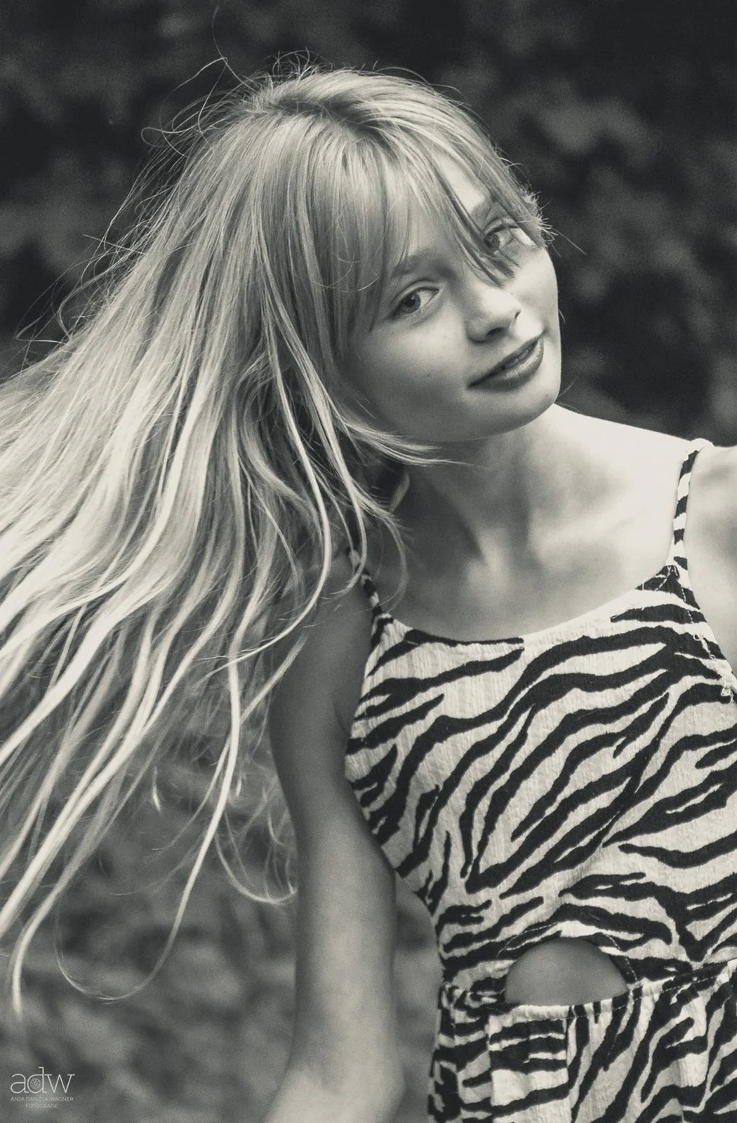 Black and white photo of a young woman with long, wavy hair, wearing a zebra-striped dress with cutouts, outdoors with blurred background.