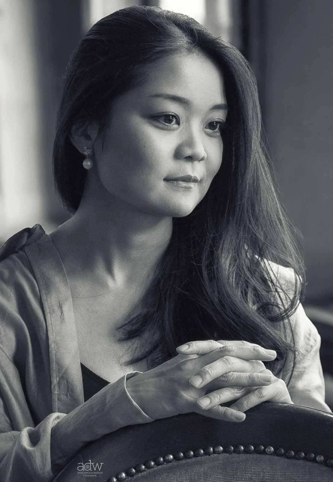 A black and white portrait of a young woman with long hair, wearing earrings, sitting with her hands clasped and looking contemplative.
