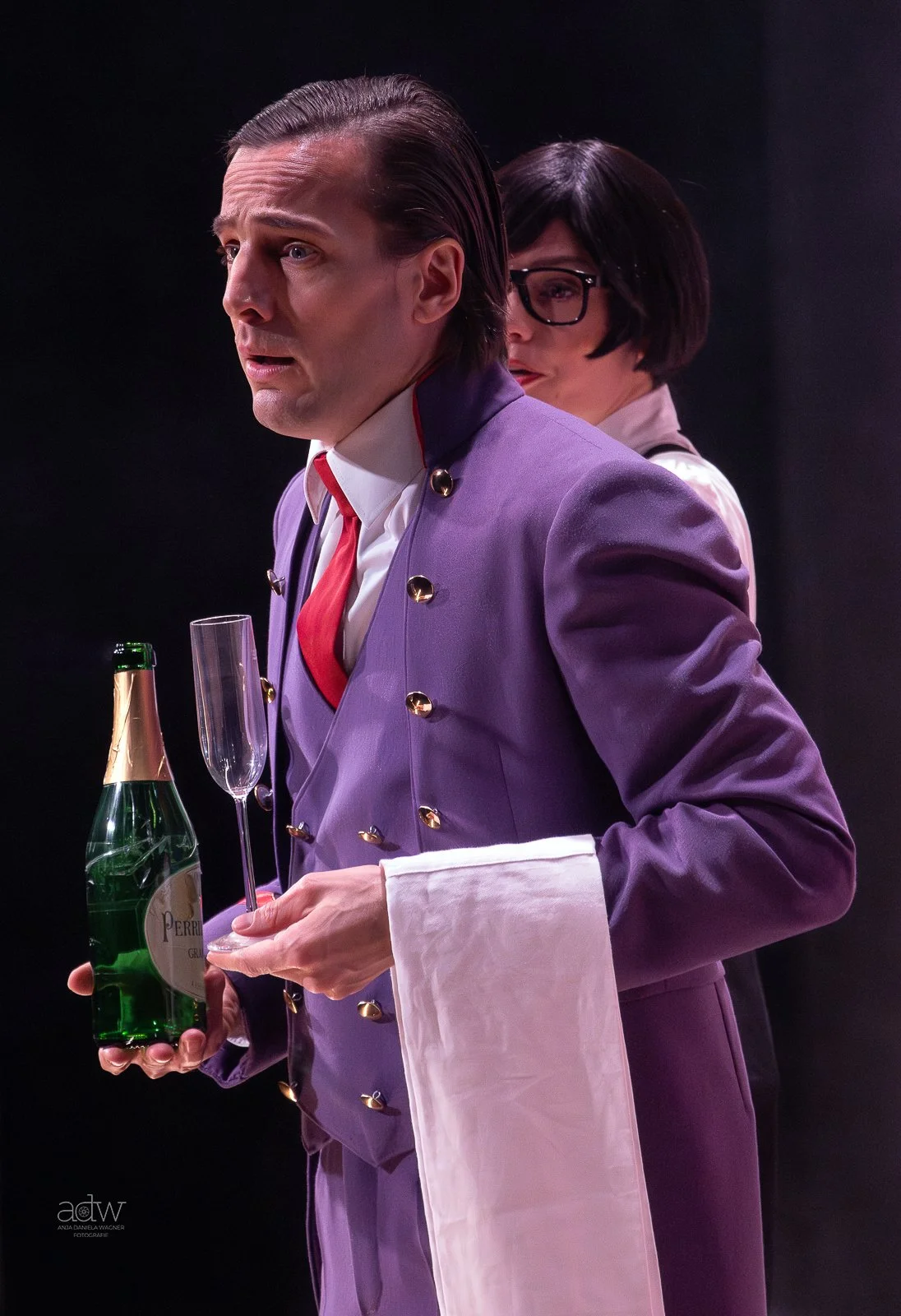 A man dressed as a royal butler or waiter holding a bottle of Perrier sparkling water and a champagne flute, with a woman standing behind him against a dark background.