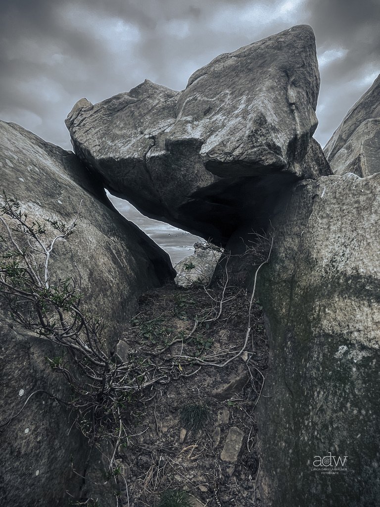 Large rocks forming a natural arch with cloudy sky overhead, small plants and tangled roots growing between rocks.