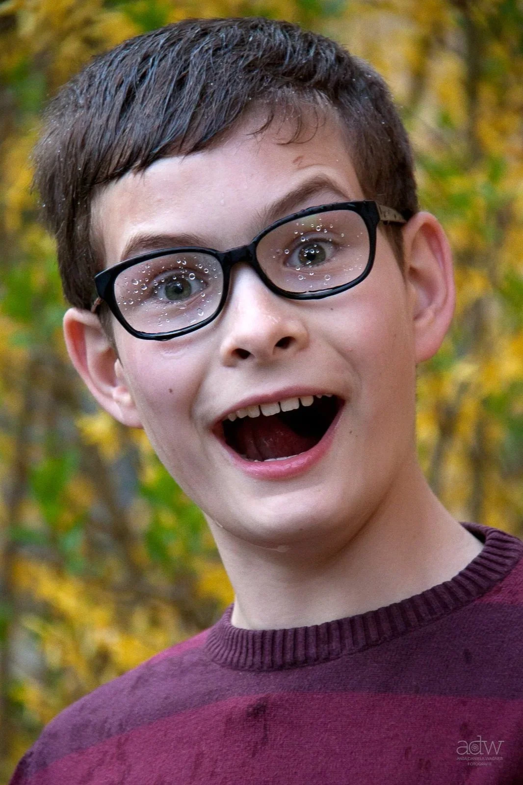 A boy with glasses smiling and looking surprised outdoors with fall-colored leaves in the background.