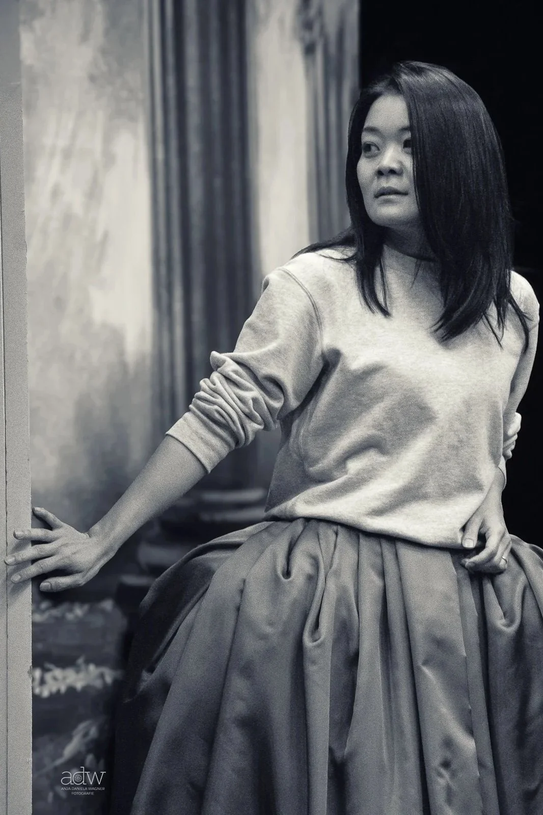 A black-and-white photo of a woman with shoulder-length dark hair, wearing a sweatshirt and a voluminous skirt, leaning against a doorway or wall, looking off to the side.